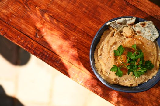 Overhead view. Chickpea hummus dip with pita bread, parsley and sprinkled paprika on a blue ceramic bowl, on a rustic wooden table background with copy ad space for text. Still life. Food composition