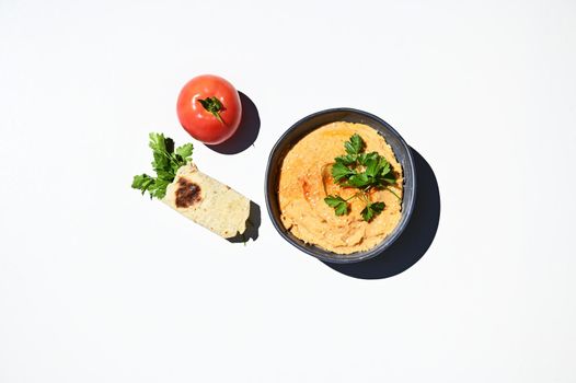 Flat lay composition of blue ceramic bowl with chickpea hummus, sprinkled with red chili, red ripe tomato and parsley wrapped in pita bread, isolated on white background with copy ad space for text