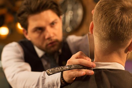 Handsome bearded barber using a razor shaving his client working at the barbershop style styling barbering hairdresser stylist professional occupation job concentration hipster lifestyle masculine.