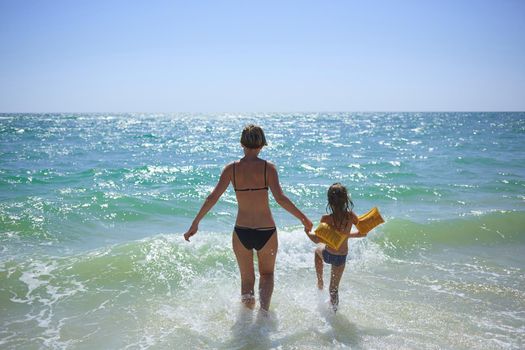 Summer happy family of six years blonde child playing and jumping water waves embracing woman mother in sea shore beach.