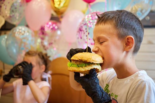 The handsome little boy eating burger in black rubber gloves