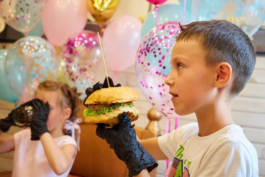 The handsome little boy eating burger in black rubber gloves