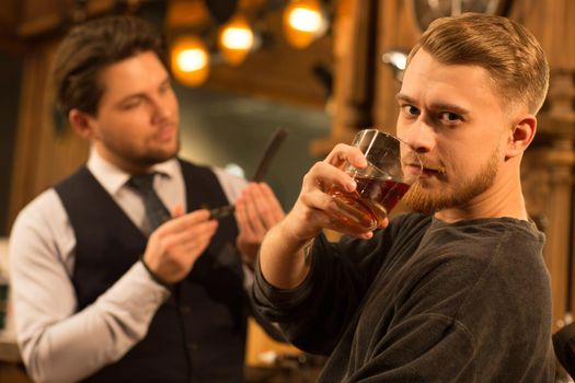 Young bearded handsome man looking to the camera fiercely drinking whiskey at the barbershop professional barber cleaning his razor on the background copyspace hipster service lifestyle masculine brutal.