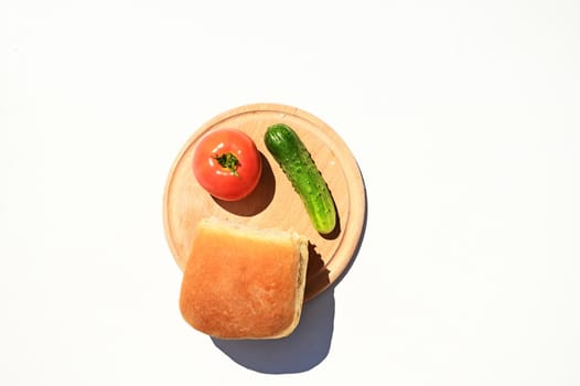 Top view of a wooden board with a loaf of freshly baked bread and raw tomato and cucumber, isolated over white background with copy space for advertising text. Food composition, still life. Flat lay