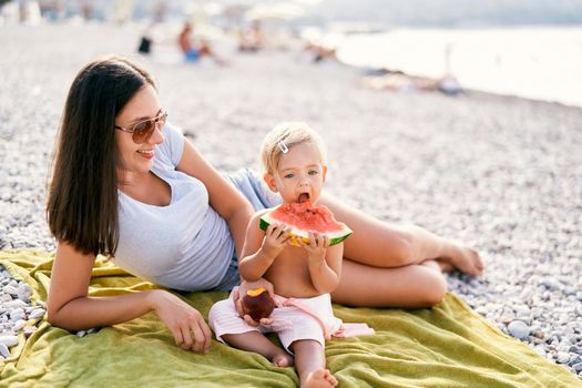 Smiling mother holding out a peach to a little girl biting a watermelon. High quality photo