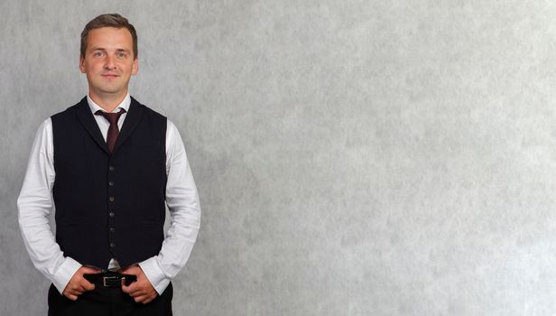 Smiling handsome man in a white shirt and grey vest looking at the camera. A man posing while holding his hands on a waist belt.