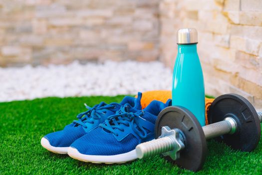 sports equipment in the foreground on green grass in the garden. orange towel, water bottle, weights and blue running shoes. stone wall background. natural outdoor light.