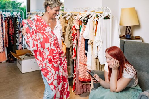 mature woman trying on clothes happy, young woman bored waiting for her friend to finish shopping for clothes. mother and daughter shopping in a clothing shop. concept of shopping. natural light from window.