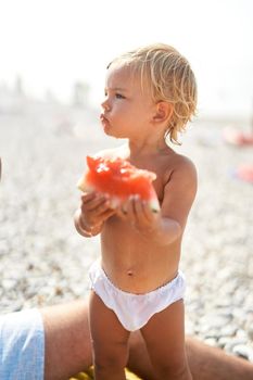 Little girl with a piece of watermelon stands on the beach and looks away. High quality photo