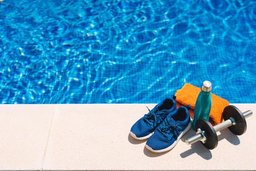 sport accessories in front of a swimming pool. orange towel, blue water bottle, weights. background of a swimming pool in a garden. outside with natural light.