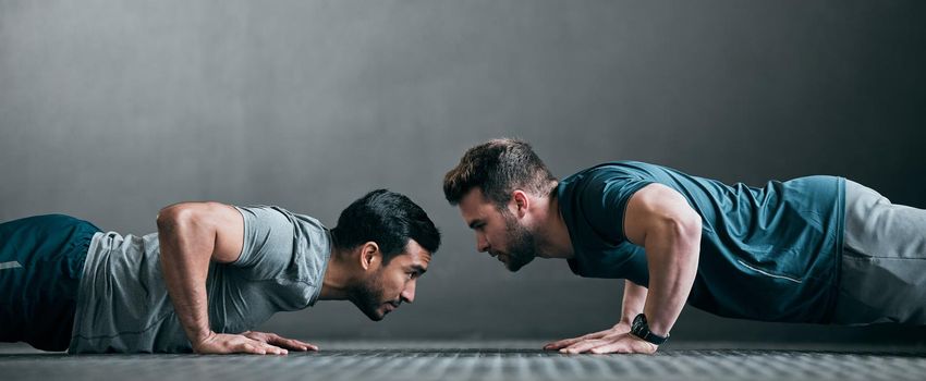 Game on. Full length shot of two handsome young male athletes doing pushups face to face against a grey background