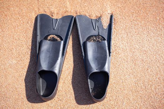 A pair of black flippers on the background of sand next to the water, top view. Swimming equipment - fins on the shore. Summer holidays, fun, exploring the sea world concept. Space for copy.