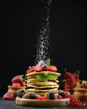 A stack of pancakes with fresh fruit sprinkled with powdered sugar on a black background