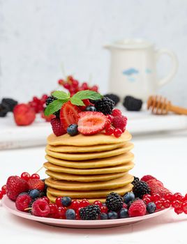 Stack of baked pancakes with fruits in a round plate on a white table, delicious breakfast