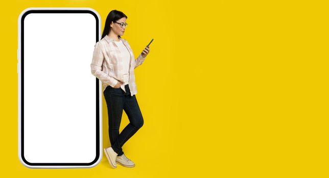 Leaned on big smartphone with white screen charming middle aged asian woman looking at phone in her hand wearing casual isolated on yellow background. Copy space on right, phone mock up.