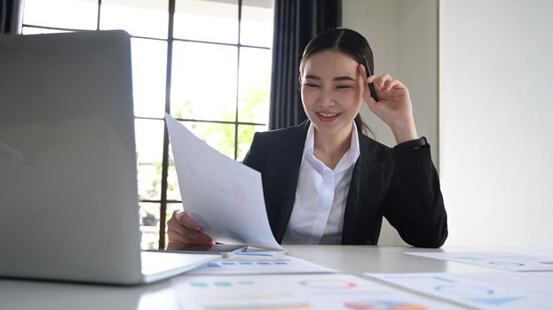 Smiling working woman using laptop computer and examining financial document at office desk.