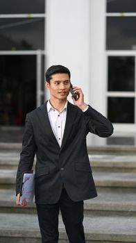 Handsome businessman in black suit standing near modern office building and talking on mobile phone.
