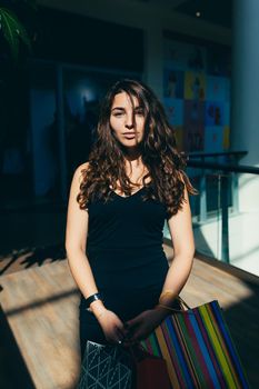 Portrait of a beautiful young woman in a black dress and curly hair in the mall who is shopping in shopping bags, looking at the camera, smiling