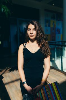 Portrait of a beautiful young woman in a black dress and curly hair in the mall who is shopping in shopping bags, looking at the camera, smiling