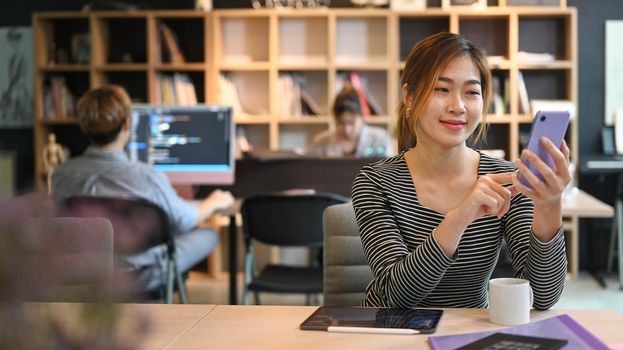 Smiling asian woman employee sitting in crate office and using mobile phone.