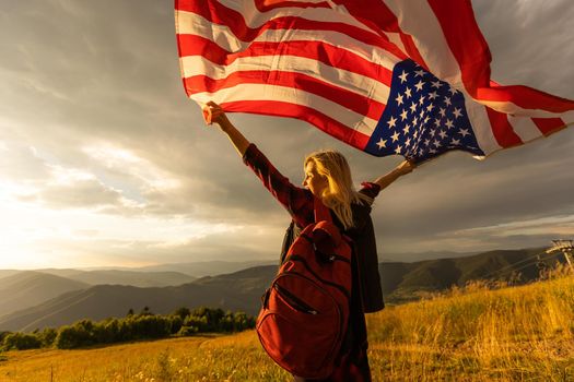 Young happy american woman with long hair holding waving on wind USA national flag on her sholders relaxing outdoors enjoying warm summer day.