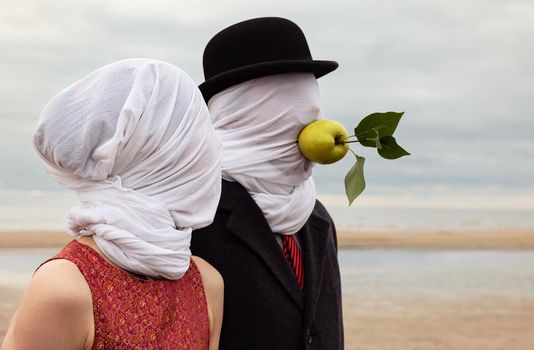 Faceless portrait of man and woman on sea background with white fabrics on their heads and green apple in a mans mouth