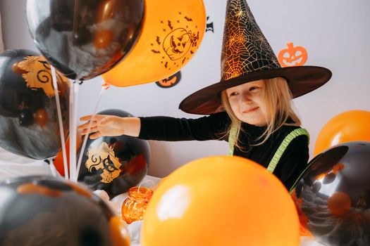 Children's Halloween - a girl in a witch hat and a carnival costume with airy orange and black balloons at home. Ready to celebrate Halloween.