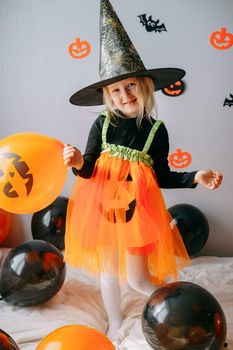 Children's Halloween - a girl in a witch hat and a carnival costume with airy orange and black balloons at home. Ready to celebrate Halloween.