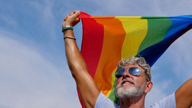 Portrait of a gray-haired elderly Caucasian man with a beard and sunglasses holding a rainbow LGBTQIA flag against a sky background. Bisexual Gay Celebrates Pride Month Coming Out Day