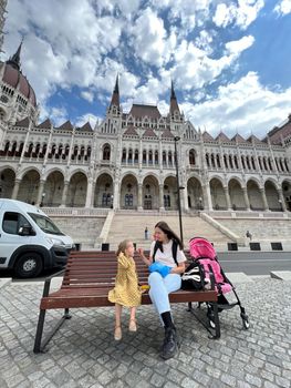 Mom and a little girl sit on a bench near the House of the Hungarian Parliament. Budapest. High quality photo