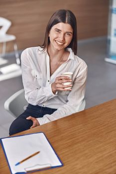 Joyful caucasian lady is drinking a coffee from a white paper cup while sitting on reception