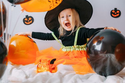 Children's Halloween - a girl in a witch hat and a carnival costume with airy orange and black balloons at home. Ready to celebrate Halloween.