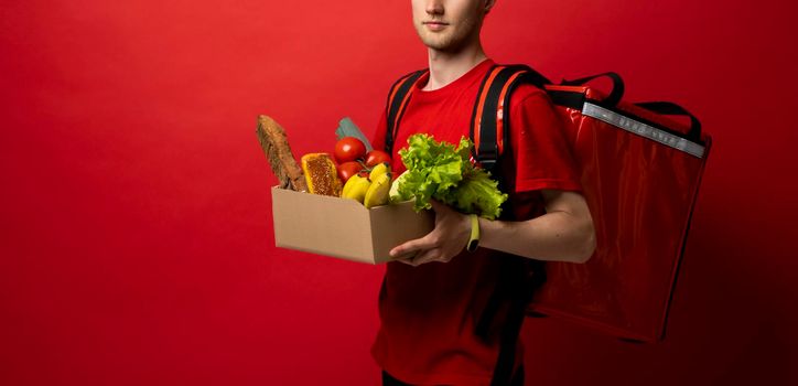 Crop picture of delivery man in red uniform carrying paper box with food products isolated over red background. Food delivery service