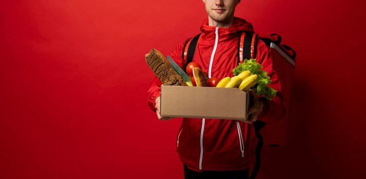 Male delivery man in red uniform holds paper box with a vegetables on red background