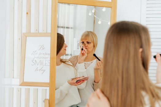 Beautiful smiling young caucasian mature senior mother mom and adult daughter doing makeup, standing near mirror, and looking at reflection in living room home. Family with two women relax together.