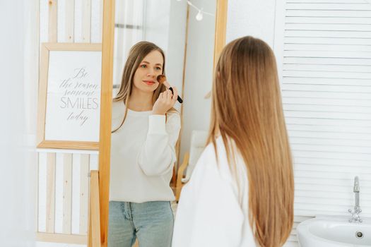 Beautiful smiling young caucasian woman with long hair standing near mirror, doing makeup and looking at reflection in living room at home.