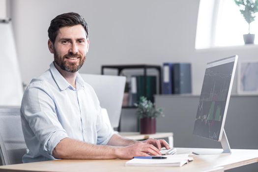 Successful bearded freelance businessman looks at camera and smiles, portrait of broker in office works at computer