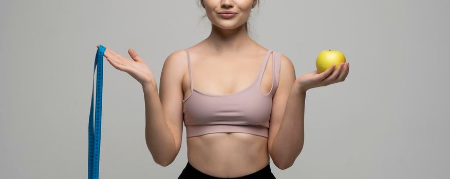 Crop image of attractive woman in a sport wear is smile and looks in a camera while holds blue measuring tape and an apple. Health care and healthy nutrition. Perfect slim body