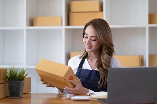 Young beautiful happy asian business woman owner of SME online using laptop receive order from customer with parcel box packaging at her startup home office, online business seller and delivery.