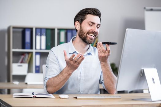 Young man with a beard, freelancer, manager, worker talking on a cell phone in the office, sitting at a computer desk, talking, waving
