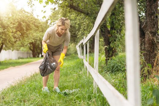 woman wearing latex gloves picking up litter to garbage bag, holding used plastic bottle in hand, takes care of planet.