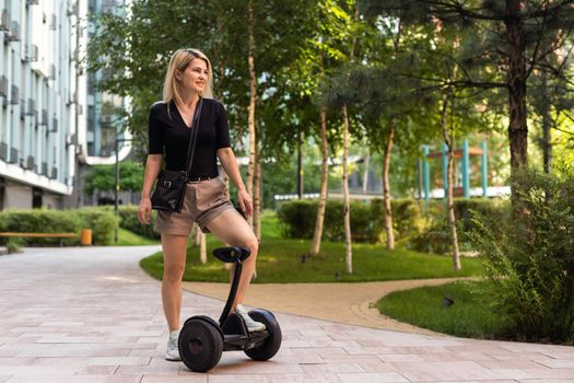 Back view of active tourist woman standing on electric self-balancing scooter at sunny park, young hipster girl driving on modern hover-board at holidays, blank space for design or content.