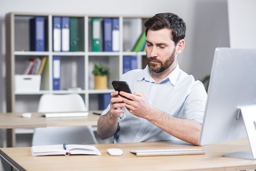 Focused young man with a beard, uses the phone, sitting at home at the table, thoughtfully reads from the screen