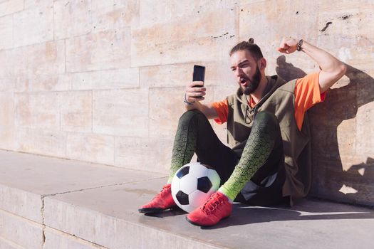 handsome soccer fan celebrates a victory when he is consulting his smart phone, concept of technology and urban sport lifestyle in the city, copy space for text