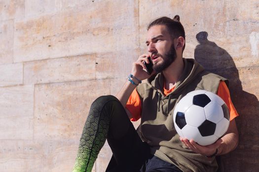 handsome sportsman talking on the phone while resting with his football ball, concept of technology and urban sport lifestyle in the city, copy space for text