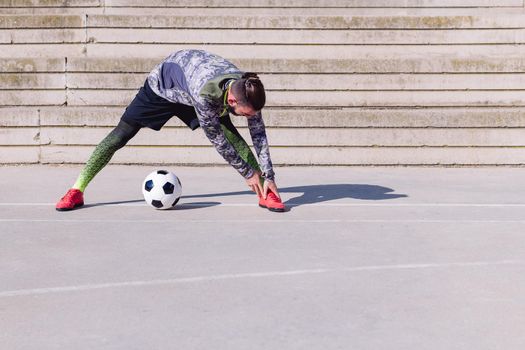 unrecognizable sportsman stretching next to his football ball in a concrete soccer court, concept of healthy lifestyle and urban sport in the city, copy space for text