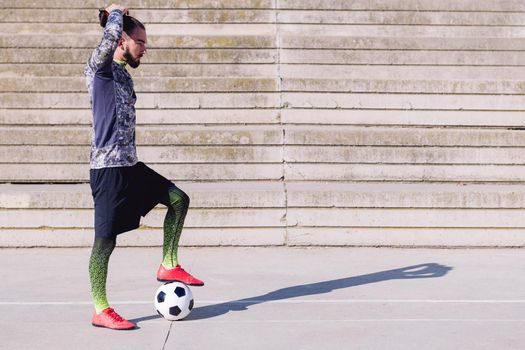 sportsman picking up his hair in a tail with a foot on his football ball in a concrete soccer court, concept of healthy lifestyle and urban sport in the city, copy space for text