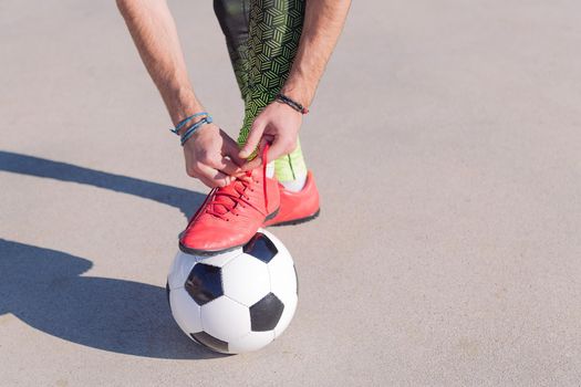 detail of a football player tying his boots with a foot on the ball in a concrete soccer field, concept of healthy lifestyle and urban sport in the city, copy space for text