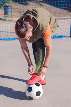 vertical photo of a football player tying his boots with a foot on the ball in a concrete soccer field, concept of healthy lifestyle and urban sport in the city, copy space for text