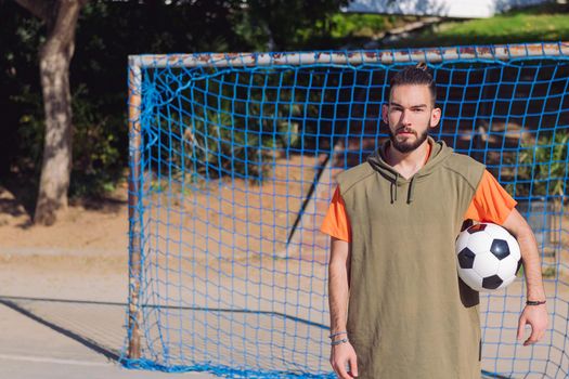 handsome hipster soccer player in front of the goal of an urban court with the ball under his arm, concept of healthy lifestyle and urban sport in the city, copy space for text